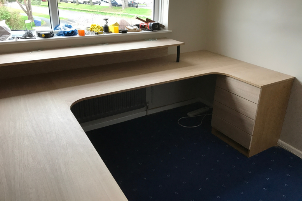 A corner desk positioned by a window, featuring a shelf above for storage or display.